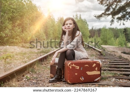 Teen girl in stylish beige dress sitting on vintage suitcase on abandoned railway in spring forest, lonely, looking at camera. Fashionable lady in retro image. Travel vacation concept. Copy text space