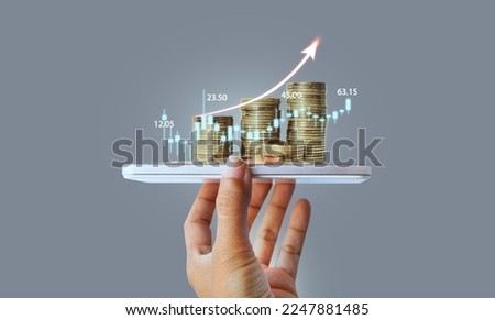 Hand holding smart phone and stack of coins over stock market screen and financial graph in background. Digital marketing hand holding smart phone with graphic icon