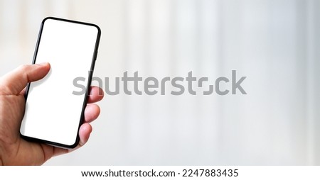 Hand holding a smartphone with blank white screen. White office background. Horizontal banner.
