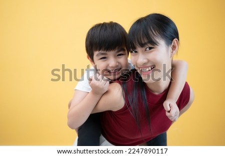 Happy young Asian woman fun  cute child baby boy 5-6-7 years old in red t-shirt stand behind hug kiss. Mommy little kid son together isolated on yellow background studio Mother's Day love family