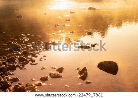 Warmy golden sunlight on quiet water surface of lake, orange haze, ripple sun glares, blinks, reflection, stones on shore in early morning, idyllic, tranquil wild nature background, texture, detail.