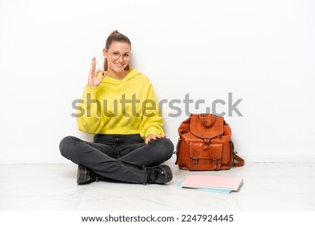 Young student caucasian woman sitting one the floor isolated on white background showing ok sign with fingers