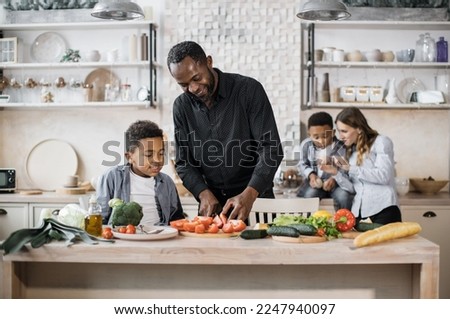 Handsome african father and son preparing tasty salad at kitchen while choping vegetables. Mom teaching smoller male kid at the background. Happy parents and children spending time together at home.