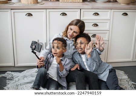 Happy multinational family of mother and two sons sitting on warm floor in light kitchen during weekend, resting and taking positive photo using selfie stick and waving hands. Royalty-Free Stock Photo #2247940159
