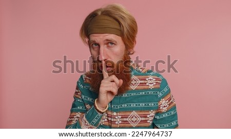 Shh be quiet please. Portrait of bearded redhead hippie man 20 years presses index finger to lips makes silence gesture sign do not tells secret. Young handsome hipster guy on pink studio background
