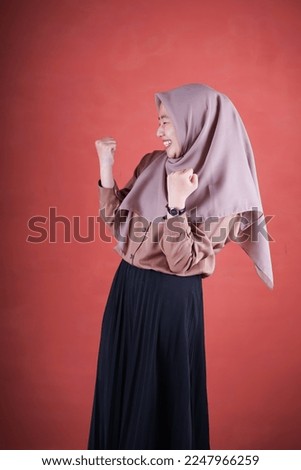              Beautiful Asian woman in brown shirt and hijab smiling cheerful winner gesture celebrate clenching fists say yes on brown background                  
