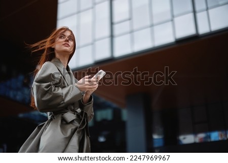 Stylish business woman with phone in hand in trendy clothes walking around town near office towers, work online technology Royalty-Free Stock Photo #2247969967