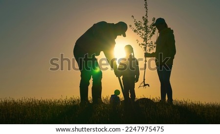 Farmer dad, mom child planting tree. Happy family team planting tree in sun spring time. Silhouette of family with tree at sunset. Family with shovel and watering can plants young trees sprout in soil