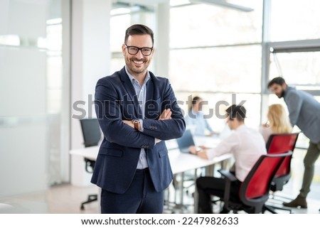 Portrait of confidence business man in formalwear standing with crossed hands at office with colleagues in the background Royalty-Free Stock Photo #2247982633