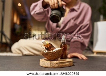 Blurred female photographer taking photo of bottles with oil and coconut on wooden board on camera at studio. Beauty and body care content for photostocks, commerce, social networks and advertising