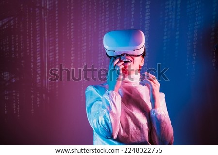 Metaverse concept. Excited, surprised woman in holographic clothes and vr glasses on matrix code background, playing video games with virtual reality headset in pink blue neon colors. Copy space.