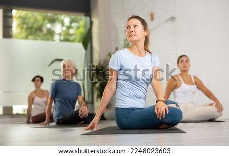 Relaxed middle-aged female performing Ardha Matsyendrasana position during group yoga training in fitness club Royalty-Free Stock Photo #2248023603
