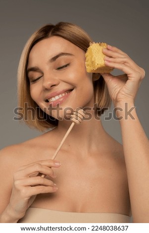 smiling woman with closed eyes holding sweet honeycomb and wooden dipper isolated on grey Royalty-Free Stock Photo #2248038637