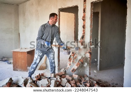 Repairman crashing brick wall with sledgehammer, pieces of brick flying away from strong blow. Destruction of partitions in flat during overhaul. Royalty-Free Stock Photo #2248047543