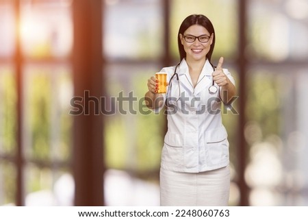 Young female doctor is showing medicine can and giving thumb up. Advertise and promotion. Blurred checkered background.