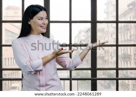 Portrait of young asian woman advertising copy space with both hands. Checkered windows with cityscape on the background.