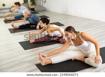 Concentrated young girl making stretching legs with group of active adult women sitting on yoga mat and doing exercise yoga together