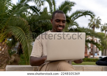 African American male programmer in casual clothes smiling and browsing laptop while sitting on border near palm trees and working remotely 