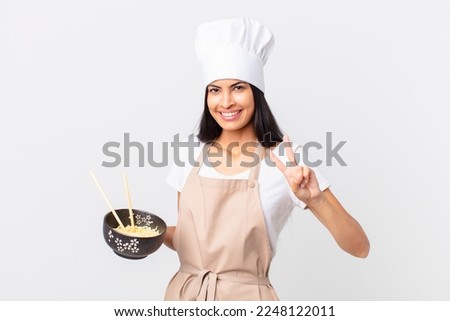 pretty hispanic chef woman smiling and looking friendly, showing number two and holding a noodle bowl