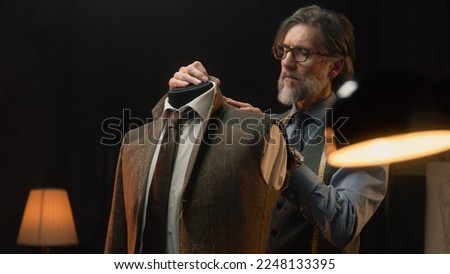 Male mature tailor works on elegant business or wedding suit. Mannequin with tailored shirt, tie and jacket in stylish luxury designer atelier or tailoring dim studio. Fashion and hand craft concept. Royalty-Free Stock Photo #2248133395