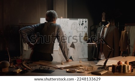 Male mature tailor works in luxury designer atelier or tailoring studio. He looks at board with sketches of future clothes and thinks. Mannequin with tailored suit. Fashion and hand craft concept. Royalty-Free Stock Photo #2248133629