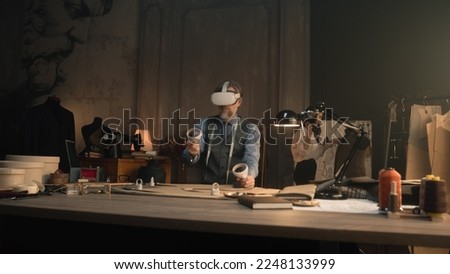 Mature tailor wearing VR headset with wireless controllers, sits at the table in luxury designer atelier, gesturing and creates design in virtual reality. Concept of technologies of future in business Royalty-Free Stock Photo #2248133999