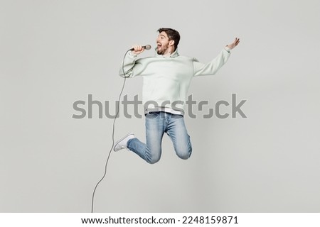 Full body young expressive singer fun man wear mint hoody jump high sing song in microphone on stage raise up hand isolated on plain solid white background studio portrait. People lifestyle concept