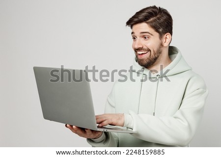 Young smiling programmer happy fun caucasian man wear mint hoody hold use work on laptop pc computer typing message isolated on plain solid white background studio portrait. People lifestyle concept