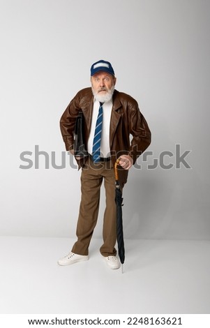 Portrait of senior old man in leather jacket posing with briefcase and umbrella over grey background. Employee. Concept of emotions, facial expression, lifestyle, modern fashion
