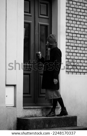 A woman on the porch of a house knocks on the door. Black and white photo.