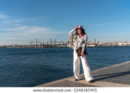 Stylish seashore woman. Fashionable woman in a hat, white trousers and a light sweater with a black pattern on the background of the sea.