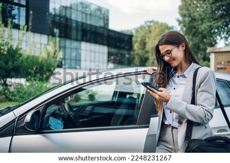 Portrait of a confident business woman wearing elegant suit and typing message on a smartphone while getting into the car. Businesswoman using mobile phone while standing next to open car door. Royalty-Free Stock Photo #2248231267