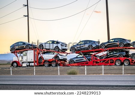 Industrial grade popular big rig white car hauler semi truck tractor transporting fastened cars on two level modular semi trailer running on the highway road at sunset in California Royalty-Free Stock Photo #2248239013