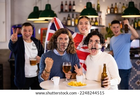 Happy young adult fans waving British flag while drinking beer and watching match together in sports bar