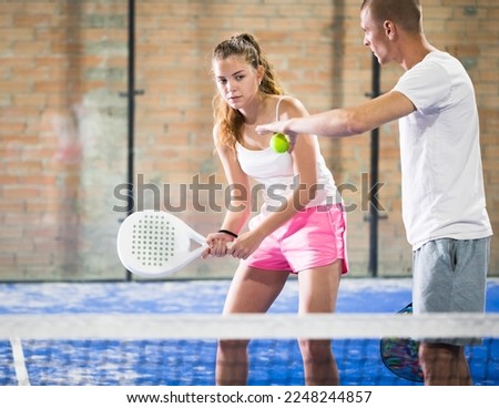 Man teaching young female padel player to serve ball while training on indoor tennis court. Focused girl swinging racket, ready to hit ball held by coach. Royalty-Free Stock Photo #2248244857