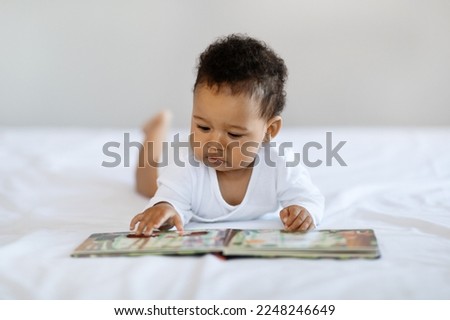 Baby Development Concept. Cute Black Infant Child Lying On Bed With Book, Adorable Little Boy Or Girl Looking At Pictures With Interest While Relaxing In bedroom At Home, Closeup Shot With Copy Space