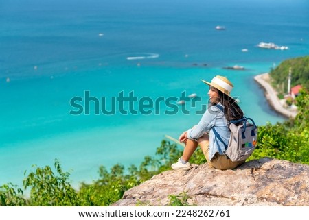 Asian woman with backpack travel at tropical island and resting on mountain peak in summer sunny day. Attractive girl enjoy outdoor lifestyle looking beautiful ocean nature on beach holiday vacation. Royalty-Free Stock Photo #2248262761