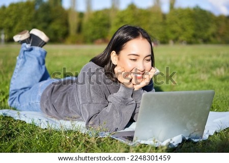 Smiling asian girl lying in park on grass, watching movie or video on laptop, looking at screen with interest.