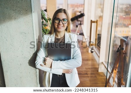 A young business woman with long blond hair in a white jacket and glasses stands with a laptop in her hands in an office space. Work in a modern office with large windows.