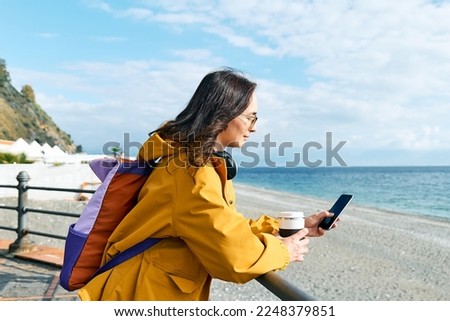 Woman with backpack using smartphone while leaning on handrail along seaside promenade. Relaxing outdoors. The concept of hiking and travel in the low season.