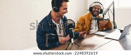 Cheerful podcasters laughing and having a good time in a home studio. Two happy young men co-hosting a live audio broadcast. Two young content creators recording an internet podcast.