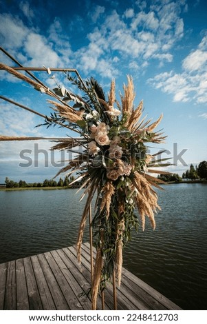 Art Deco wedding arch with pink roses and leaves on a wooden platform for the ceremony, against the background of the river, on a clear sunny day.