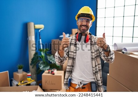 Young hispanic man with beard working at home renovation success sign doing positive gesture with hand, thumbs up smiling and happy. cheerful expression and winner gesture. 