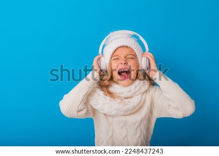 portrait of a happy little blonde girl in a white hat and sweater listening to music with headphones on a blue background. Winter, space for text. High quality photo