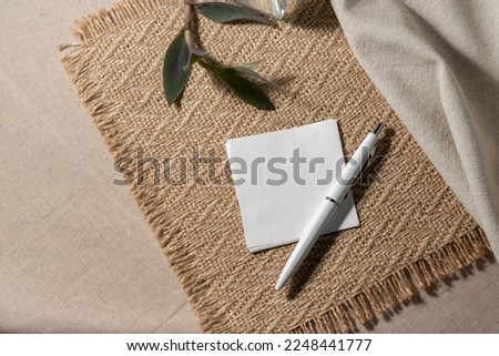Blank paper, invitation card sheet with empty mock up copy space, pen and plant on a neutral beige jute placemat and linen cloth