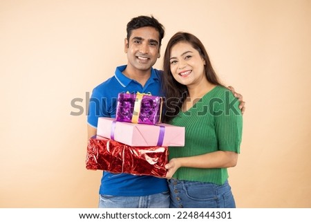 Portrait of happy indian couple wearing casual T-shirt holding Gift boxes and celebrating festival together isolated on beige background. Boy give present to girl, Valentines day celebration.