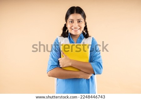 Happy Rural Indian student schoolgirl wearing blue government school uniform holding books and bag standing isolated over beige background, Studio shot,closeup, Education concept.