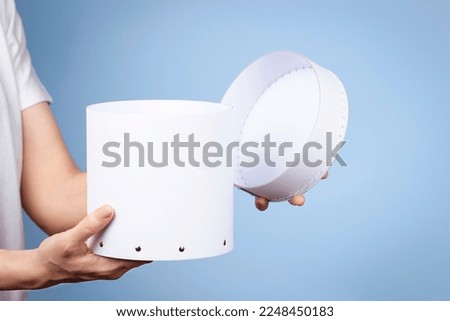 Woman hands holding white round cardboard box and open cap on light blue background. Mock up package box