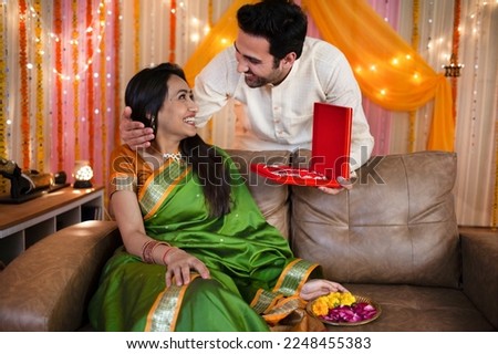 Potrait of a young romantic couple with gift box on festival or birthday or anniversary. Stock image of a happy man giving a gift or necklace to his pretty wife sitting on a sofa with a plate of fl...