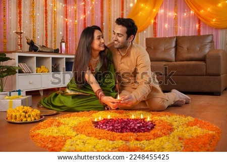 Portrait of an Indian couple looking at each other placing diyas on rangoli for Diwali - Festival celebration. Image of a happy husband placing diyas with his gorgeous wife and making flower rangol...